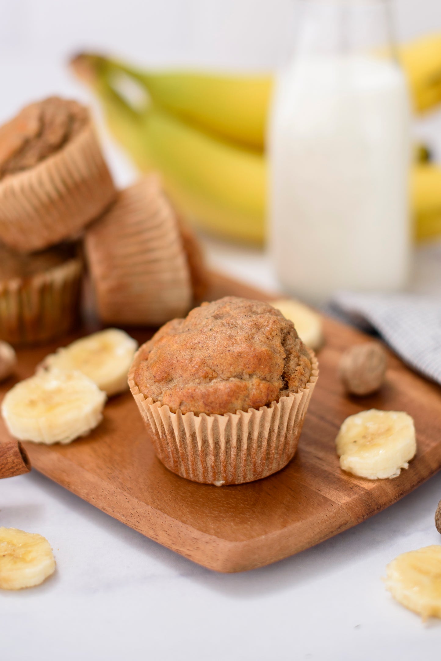 A stack of banana muffins on a wooden board with ripe bananas and a bottle of milk in the background.