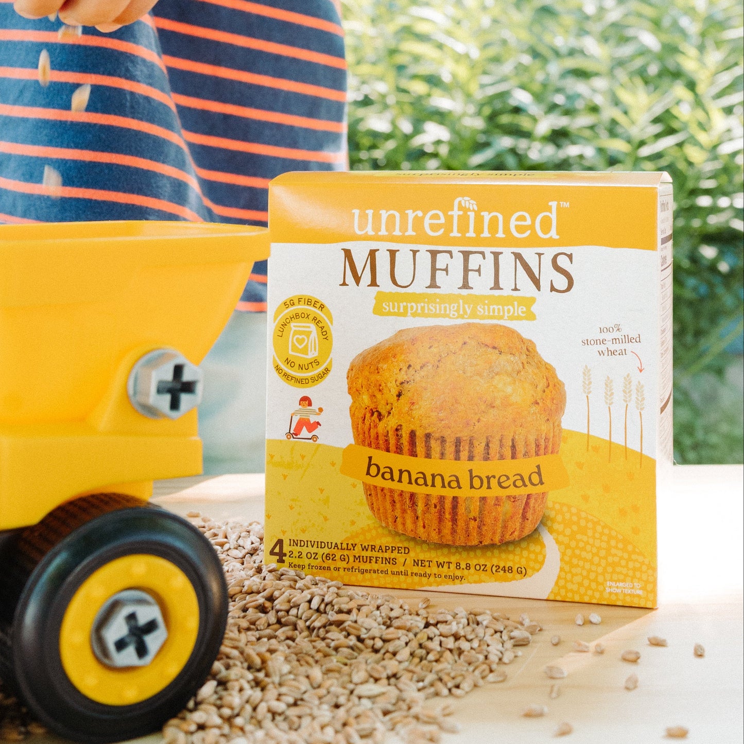 A box of Unrefined Muffins banana bread is displayed on a table outdoors with wheat seeds, a yellow toy dump truck, and a child's hand and striped shirt in the background.