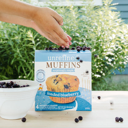 A hand reaches toward a box of unrefined 'loaded blueberry' muffins placed on a wooden table outdoors, with a bowl of blueberries and greenery in the background.