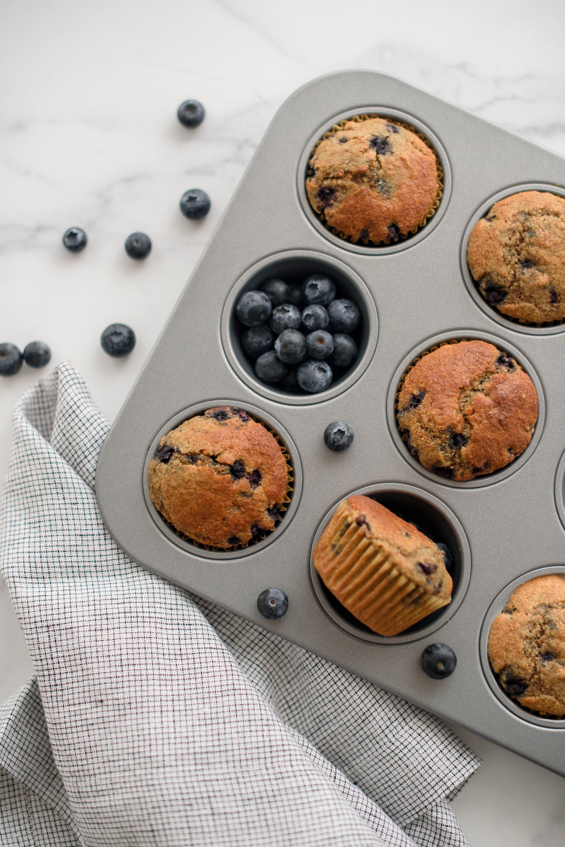 Muffin tin with blueberry muffins and fresh blueberries on a marble surface