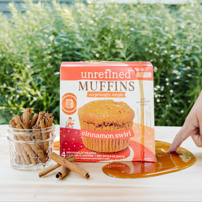 A box of Unrefined Muffins Cinnamon Swirl is displayed on a light wood table outdoors, with a hand pointing toward the product. A glass jar of cinnamon sticks and loose cinnamon sticks sit to the left, and a pool of maple syrup is in the foreground.