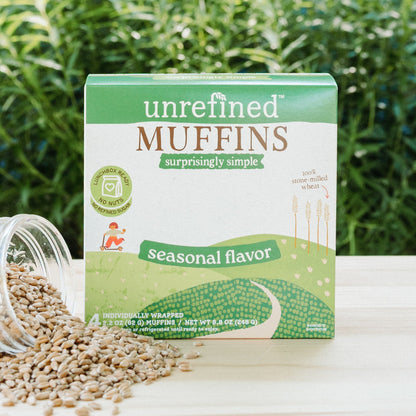 A box of Unrefined Muffins in seasonal flavor sits on a light wood surface with loose grain spilled from a glass jar beside it, set against a blurred green outdoor background.
