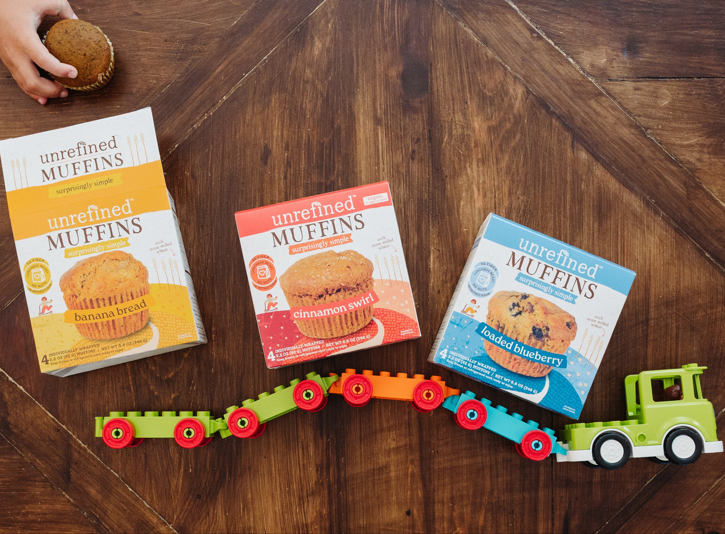 Three boxes of Unrefined Muffins arranged on a dark wood table with a colorful toy train and a child's hand holding a muffin at the top edge.