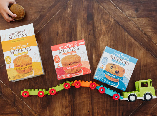 Three boxes of Unrefined Muffins arranged on a dark wood table with a colorful toy train and a child's hand holding a muffin at the top edge.