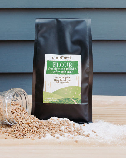 A black bag of unrefined flour stands on a light wood surface with spilled whole grains and whole grain flour in front of a blue-gray wooden plank background.