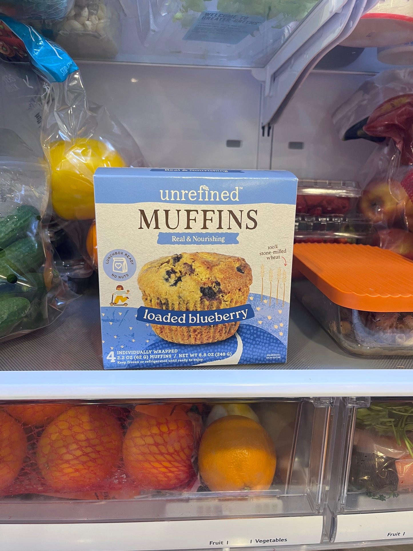 Box of blueberry muffins in a grocery store produce section with fruits and vegetables.