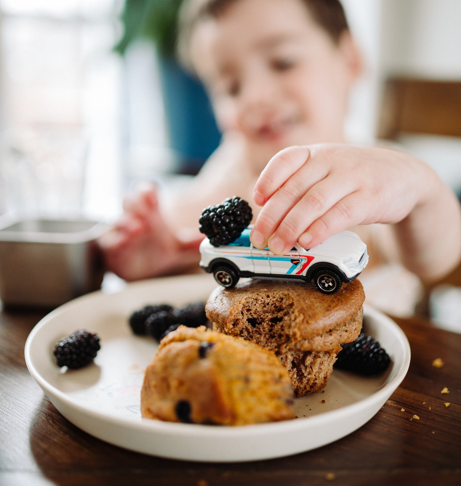 Child playing with a toy car on a muffin with blackberries on a plate.